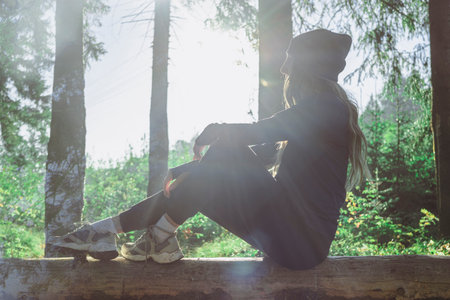 A female tourist in the summer northern forest and resting on a log. The silhouette of a woman in sportswear and a hat with a side view is sitting in the rays of the sun.の写真素材