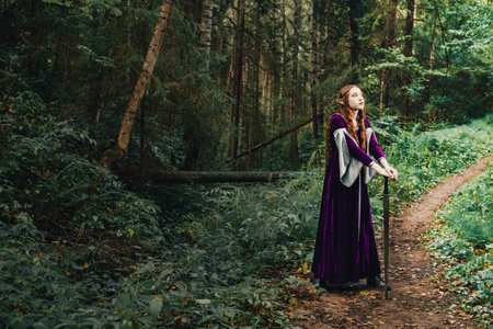 An elven girl driver with long red hair in a purple dress in the forest poses standing with a medieval sword. The woman is a fairy tale elf character.の写真素材