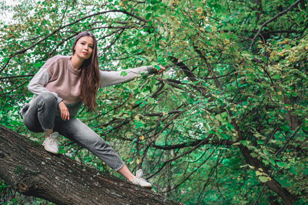 A brown haired girl in a sweater and jeans touches a branch with green summer leaves. The concept of unity with nature.の写真素材