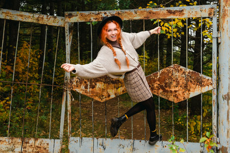 A girl in a black hat, skirt and sweater is hanging and swinging on the rusty gate of an abandoned...