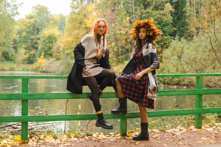 Two girlfriends in the autumn park are sitting on a green fence by the pond. Two girls laugh and communicate with each other.の写真素材