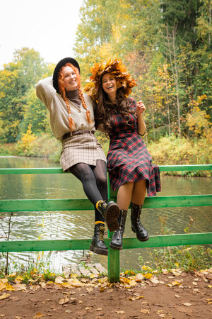 Two girlfriends in the autumn park are sitting on a green fence by the pond. Two girls laugh and communicate with each other.の写真素材