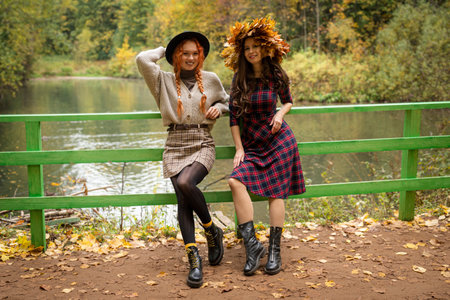 Two girlfriends in the autumn park are leaning on a green fence by the pond. Two girls laugh and communicate with each other.の写真素材