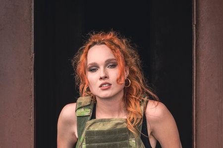 A girl in a black bodysuit with a camouflage bulletproof vest stands against an iron industrial background. Portrait aggressive red haired girl poses next to a rusty bunker.の写真素材