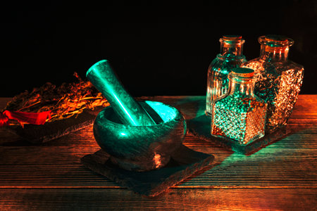 A stone mortar and pestle stand on a wooden table with glass vials and dried flowers. Magic tools in...