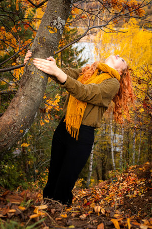 A young red haired woman hanging a tree trunk in an autumn park.の写真素材