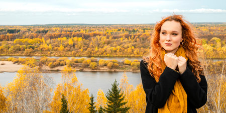 A woman in a black coat and an orange scarf stands on the high bank of an autumn river. A girl in warm classic clothes for cool weather.の写真素材