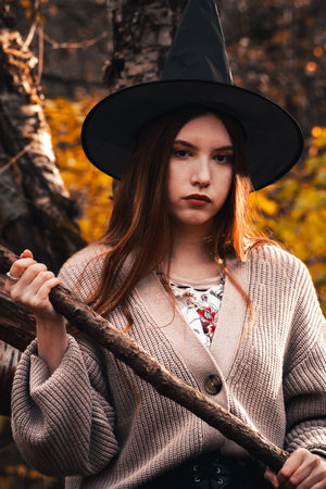 A witch woman with a broom in a black hat is sitting on a tree in the autumn forest. Halloween concept.の写真素材