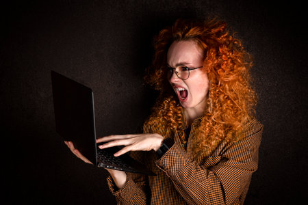 A red-haired woman in a brown shirt screams into a laptop monitor on a black dark background. The concept of frustration and aggression.の写真素材