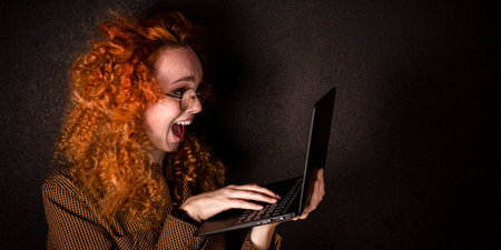 A happy red haired woman smiles and holds a laptop in her hands on a black dark background. The...