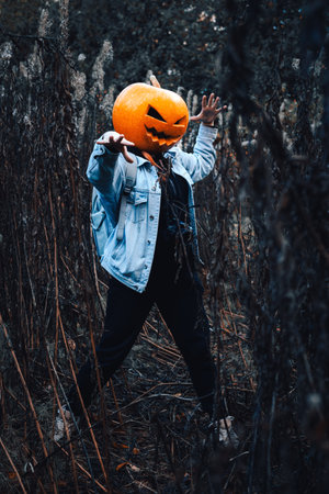 Woman with an orange pumpkin on his head in a jeans jacket in the fashion of a scarecrow stands on the outdoor. Female intimates with raised hands. The concept of celebrating Halloween.の写真素材