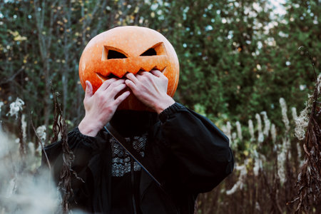 A man with an orange pumpkin on his head in a black jacket in the image of a scarecrow nail biting. The concept of celebrating Halloween.の写真素材
