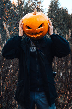 Man with an orange pumpkin (jack o lantern) on his head in a black jacket stands in the autumn dry...