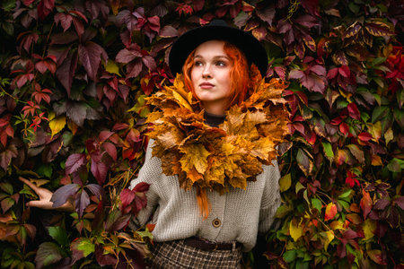Portrait of a red haired young woman with freckles in a wreath of yellow maple leaf in a gray cardigan against a red wall of creeping vine leaves.の写真素材