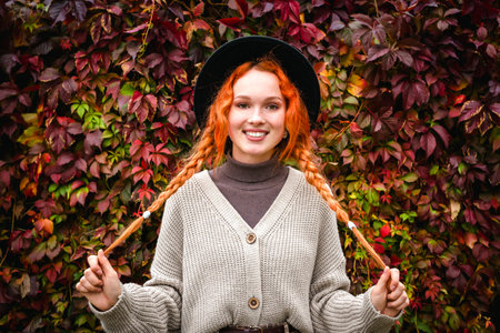 Female a smiling red haired with freckles in a black hat and a gray cardigan against a red wall of creeping vine leaves. Autumn portrait young woman.の写真素材
