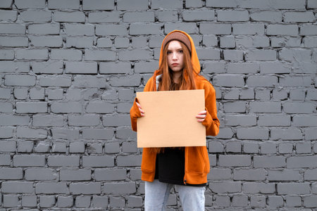 A female activist in an orange hoodie holds an empty (copy space) poster against a gray brick wall. The concept of a protest or picket.の写真素材