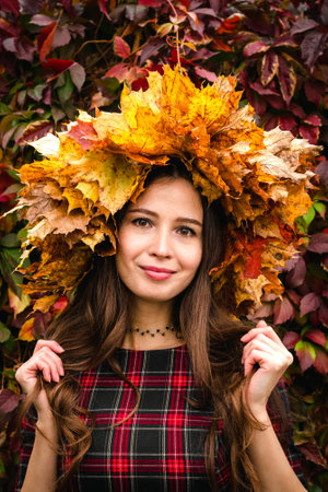 Portrait of a brown haired happy woman in a wreath of yellow maple leaves, in a plaid dress against a red wall of creeping vine leaves.の写真素材