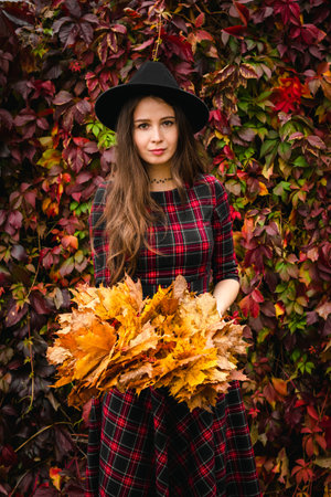 Brown haired happy woman in a black hat hold an hand is wreath of yellow maple leaves, standing in a plaid dress against a red wall of creeping vine leaves.の写真素材