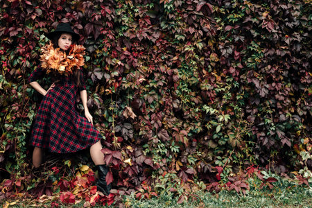 A brown haired woman in a black hat with a wreath of yellow maple leaves around her neck, stands in a plaid dress against a red wall of creeping vine leaves.の写真素材