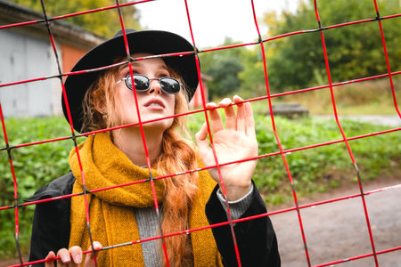 A red haired woman in a black hat and an orange scarf is holding onto a red grille in the...