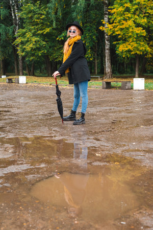 Red haired woman in black glasses and a black hat in an orange scarf on the background of a park in early autumn. The reflection of a girl in a muddy puddle in a pit on an asphalt road.の写真素材