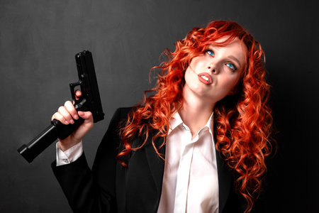 Portrait of a young red-haired woman armed with a pistol in a business suit. The concept of the business mafia.の写真素材
