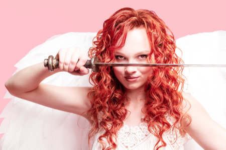 Red haired girl with white angel wings in a dress holds a medieval sword on a pink background. Goddess with a weapon. The concept of a female cupid.の写真素材