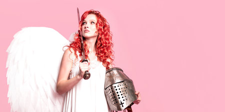 Red haired girl with white angel wings in a dress holds a medieval sword and armor helmet on a pink background. Goddess with a weapon. The concept of a female cupid.の写真素材