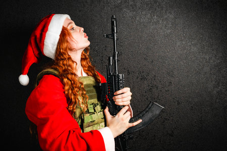 Woman in a Santa Claus costume and a red hat holds an assault rifle in her hands. Portrait of a girl in an army bulletproof vest on a black dark background. Concept of military Christmas, New Year.の写真素材