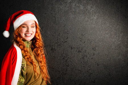 A red-haired woman in a Santa Claus costume and a red hat. Happy girl in a military army uniform on a black dark background. Concept of Christmas or New Year.の写真素材