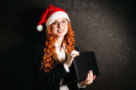 Portrait of a red-haired business woman in a suit with a laptop in a red Christmas hat.の写真素材
