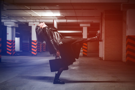 A brunette woman in a black leather jacket, dress and tights poses with a suitcase in an underground...