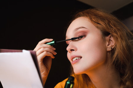 A serious and focused young woman applies makeup while looking in the mirror at home. The concept of...