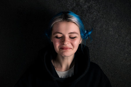 Portrait of a sad woman in a black hoodies with blue hair on a dark background