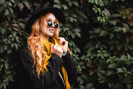 Stylish red haired woman in a black hat, glasses and a yellow scarf stands against a wall of grape...