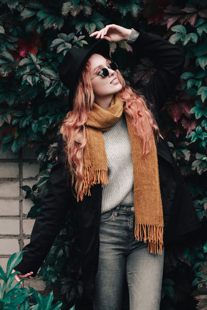 Stylish red haired woman in a black hat, glasses and a yellow scarf stands against a wall of grape leaves. Autumn portrait of a young girl.の写真素材