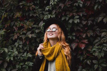 Stylish red haired woman in a black hat, glasses and a yellow scarf stands against a wall of grape leaves. Autumn portrait of a young girl.の写真素材