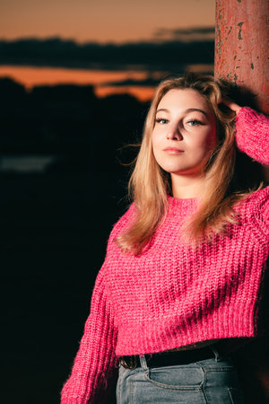 Beautiful woman on the riverbank in a pink knitted sweater poses on the background of sunset over the city landscape. Evening photo with flash.の写真素材