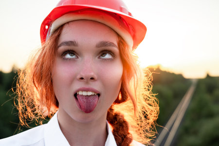 Portrait making faces woman construction inspector in a white shirt and a red hard hat on the background of a bridge being built.の写真素材