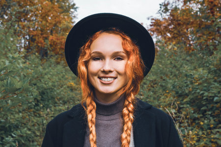 Fashionable women's autumn portrait in the park area. Red haired girl in a black hat with pigtails in a black coat stands against the background of withering foliage.の写真素材