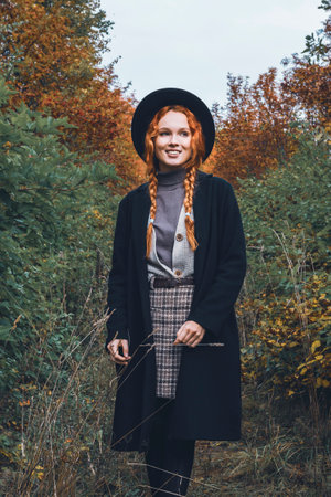Fashionable women's autumn portrait in the park area. Red haired girl in a black hat with pigtails in a black coat stands against the background of withering foliage.の写真素材