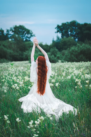 A red-haired sensual woman in a white dress of the 19th century on a background of a green field. The concept of lightness and freedom. The bride is outdoors in the summer.の写真素材
