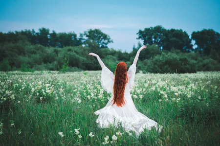 A red-haired sensual woman in a white dress of the 19th century on a background of a green field. The concept of lightness and freedom. The bride is outdoors in the summer.の写真素材