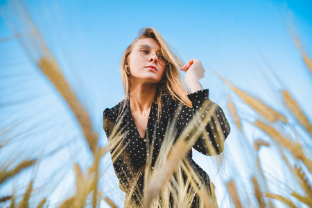A beautiful and sensual blonde woman in a black dress with white polka dot stands in a field of golden rye with a view from below against the blue sky. The concept of freedom and fertility.の写真素材