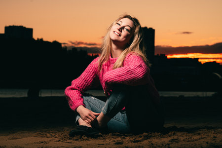 Beautiful woman is sitting on the riverbank in a pink knitted sweater against the backdrop of a sunset over an urban landscape. Evening photo with flash.の写真素材