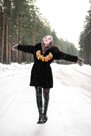 Woman in a black fur coat in a headscarf in leather boots walks along the road in a winter coniferous forest. Girl with traditional bagels on a rope.の写真素材