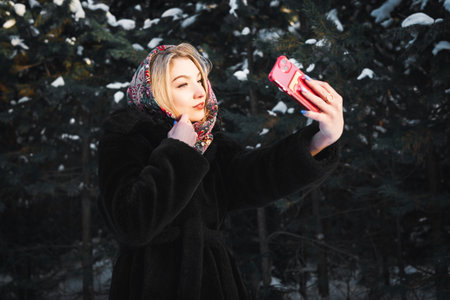 Woman in a black fur coat and a headscarf takes a selfie in a snowy winter forest against the background of fir trees.の写真素材