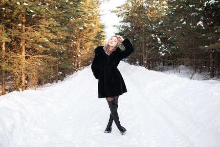 Woman in a black fur coat in a headscarf in leather boots walks along the road in a winter coniferous forest.の写真素材