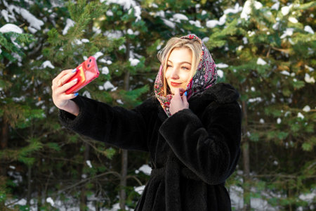 Woman in a black fur coat and a headscarf takes a selfie in a snowy winter forest against the background of fir trees.の写真素材