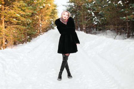 Woman in a black fur coat in a headscarf in leather boots walks along the road in a winter coniferous forest.の写真素材